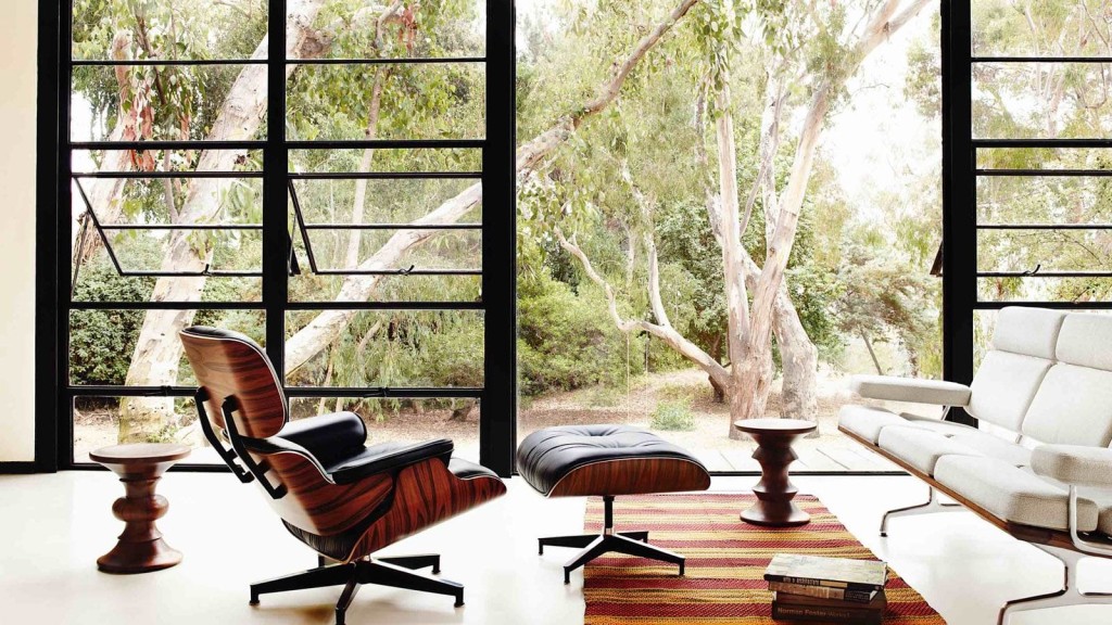 Spacious modern living room with floor-to-ceiling windows offering a view of a serene grove. Features include a mid-century modern Eames lounge chair with ottoman, a sleek white sectional sofa, and a rustic woven area rug. The room’s aesthetic is enhanced by natural light and a connection to the outdoor environment.