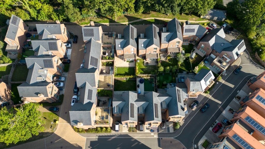 Aerial view of a newly developed residential neighbourhood featuring modern homes with solar panels on the roofs. The layout includes well-organised houses surrounding a central green space, driveways, and neatly landscaped gardens, showcasing efficient use of space and sustainable design elements.