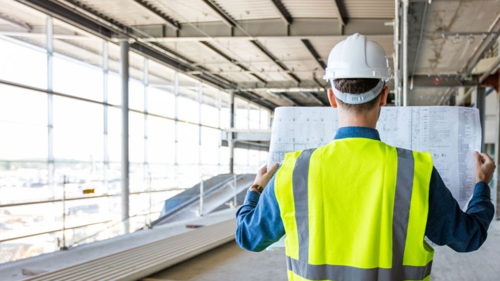 Construction worker wearing a hard hat and high-visibility vest, examining architectural blueprints inside a large, unfinished building. The image highlights the planning and inspection processes necessary for ensuring new construction projects meet minimum space standards and safety regulations.