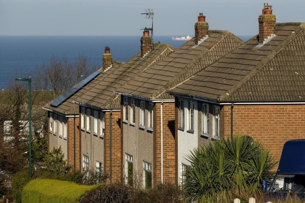 Row of traditional semi-detached houses with brick facades and tiled roofs, one featuring solar panels. The homes are set against a backdrop of the ocean, with a large ship visible in the distance, illustrating a coastal residential area adhering to modern energy standards.