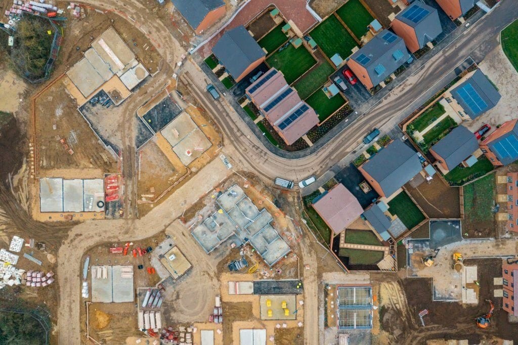 Aerial view of a residential construction site with various stages of development, including foundations and completed houses. The image shows the layout of new homes with solar panels and landscaped yards, emphasising the planning and construction process required to meet modern housing standards.