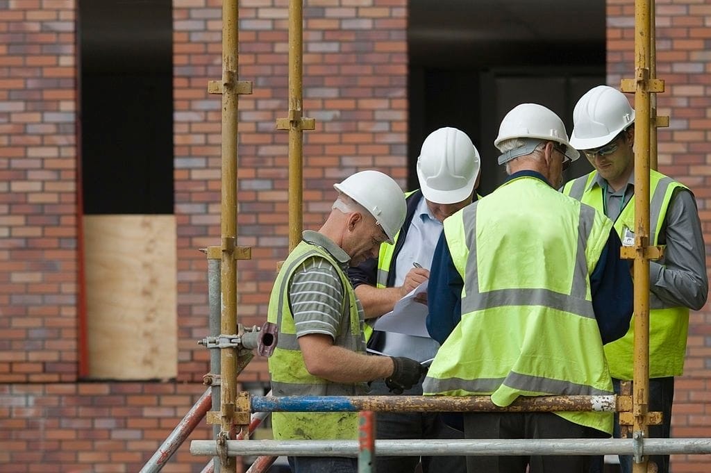 Construction workers in safety gear and hard hats engaged in a discussion on a scaffold, with a brick building under construction in the background.