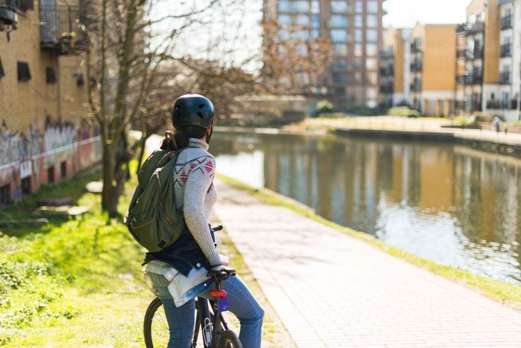 Cyclist wearing a helmet and backpack pausing by a serene canal in London, with modern residential buildings in the background and early spring foliage, embodying sustainable urban transport.