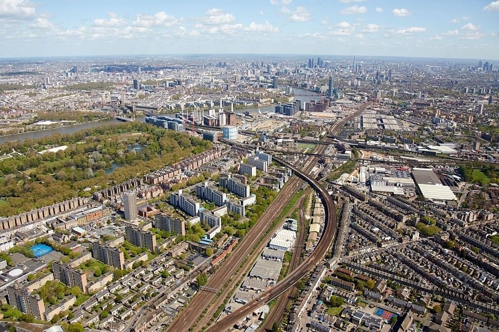Aerial view of London showcasing the city's expansive layout, with a blend of green parks, residential areas, busy train tracks, and distant cityscape under a clear blue sky.