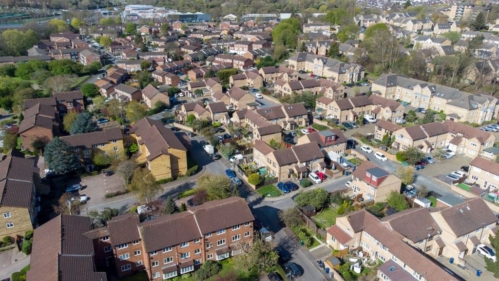 Aerial view of a suburban neighborhood in London with rows of detached and semi-detached houses, their gardens, and cars parked along the curving streets on a sunny day.