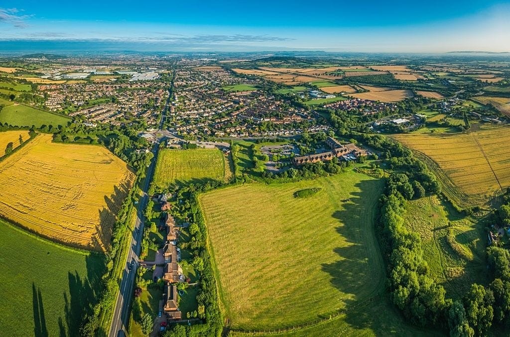 Aerial view of a lush British suburb with a patchwork of fields, homes, and a main road, showcasing the harmonious blend of residential areas and green landscapes on a clear sunny day.