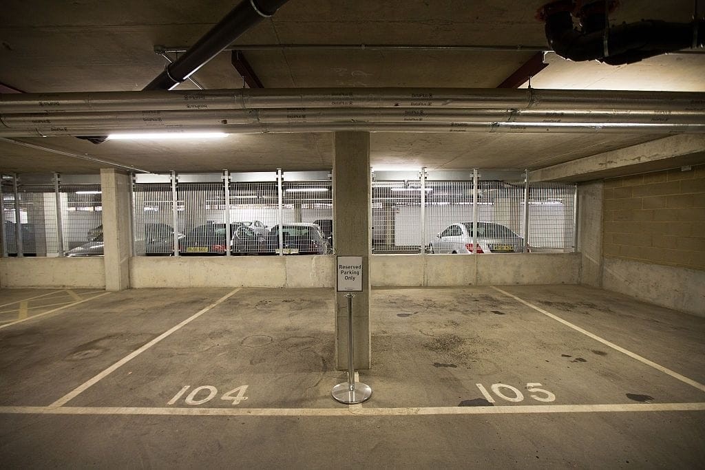 Empty parking spaces marked 104 and 105 in a well-lit underground car park, with a sign indicating 'Reserved Parking Only' and a background of parked cars behind a metal fence.