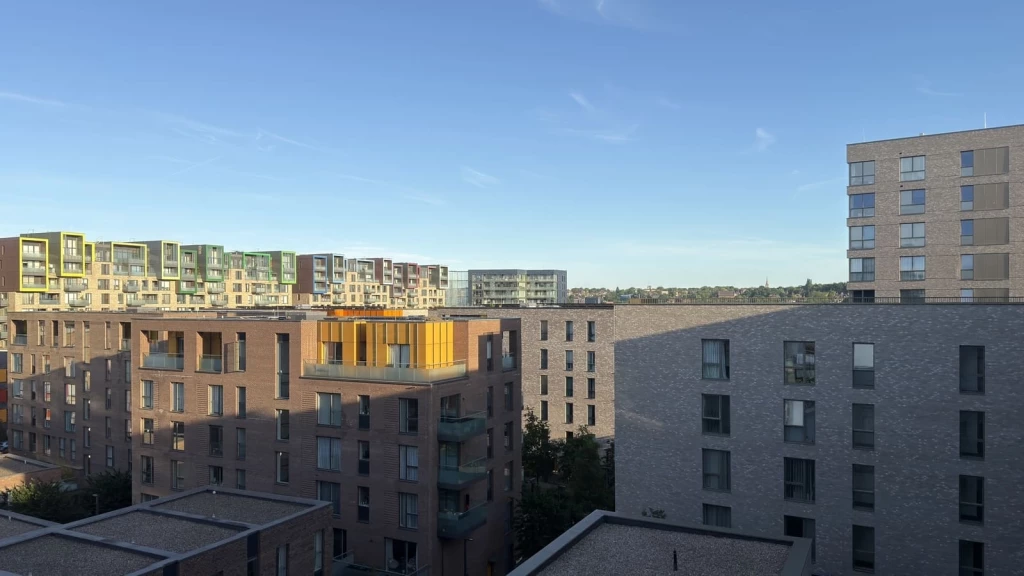A wide view of contemporary London apartment buildings designed in the New London Vernacular style, featuring brick façades, flat elevations, deep-set portrait windows, recessed balconies and muted cladding details, with a clear blue sky above and neighbouring residential blocks visible across the urban skyline.