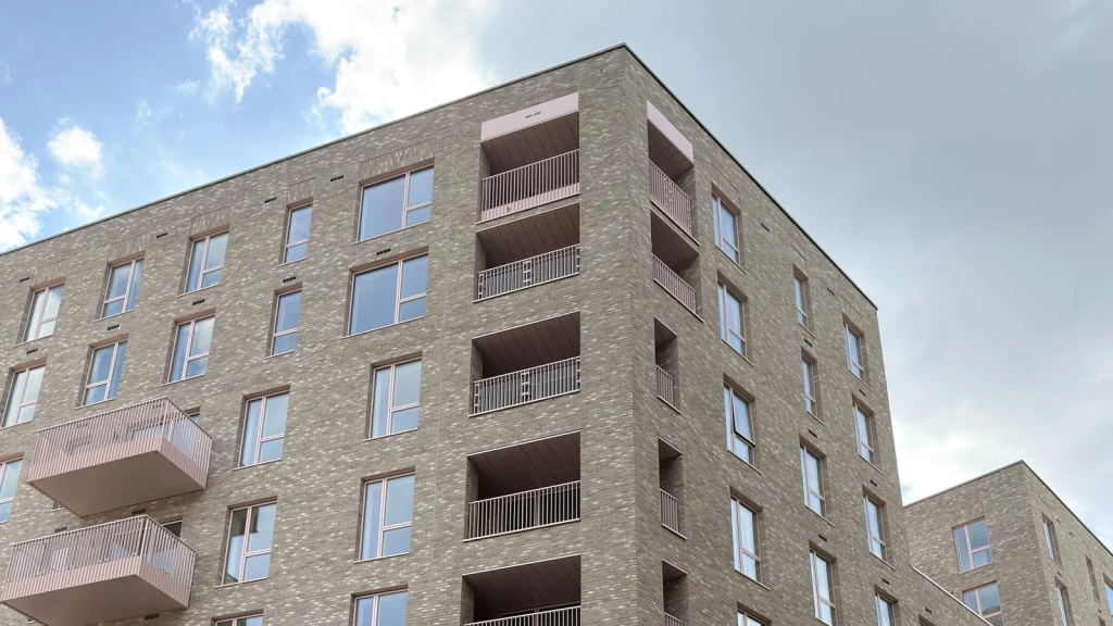A close-up view of a modern London residential building in the New London Vernacular style, showing a grey brick façade with flat elevations, deep-set rectangular windows, recessed balconies, slim metal railings and restrained contemporary detailing against a bright sky.