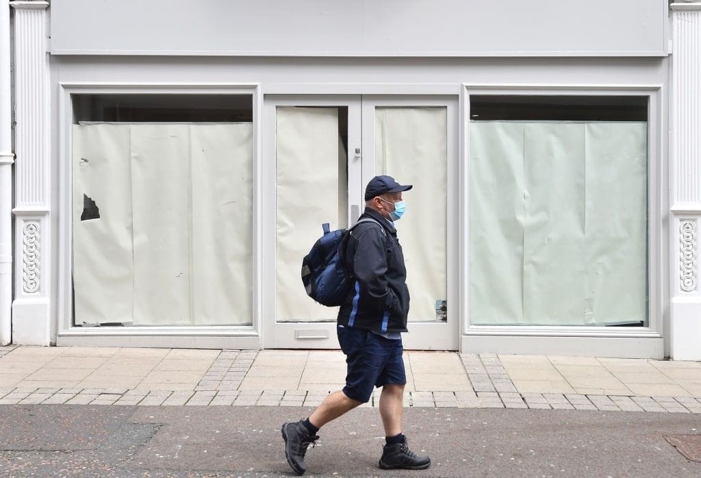 Pedestrian wearing a mask walks past a closed retail store with boarded-up windows, representing the decline of high street businesses and the effects of new Permitted Development Rights.