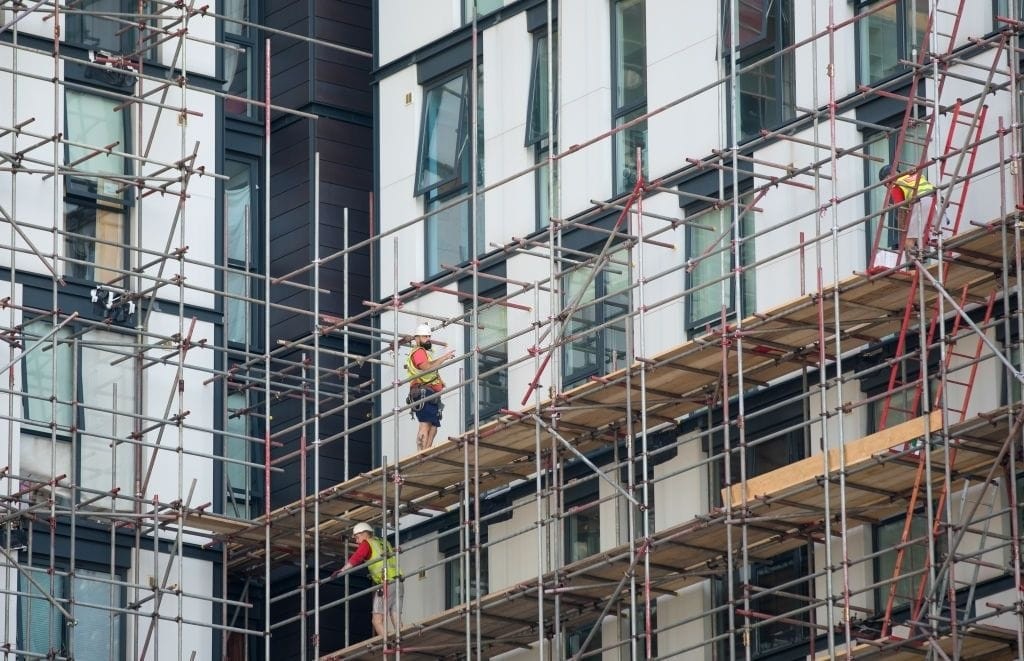 Close-up of construction workers in safety gear standing on scaffolding, adjusting and installing materials on the facade of a modern building, demonstrating the intricate work involved in large-scale urban development projects.