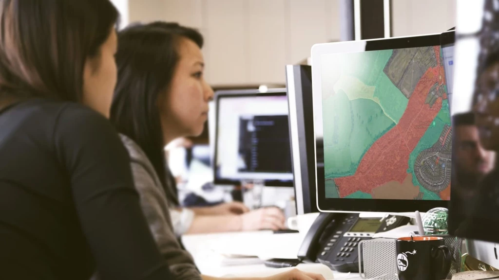 Two planning officers analyse a digital map on a computer screen, highlighting land use areas in red and green. The image is focused on assessing housing development potential and Green Belt land boundaries, key elements in urban planning. This represents the critical process of councils reviewing their Five-Year Housing Land Supply (5YHLS) data to ensure adequate land is available for housing development, in accordance with national planning policies.