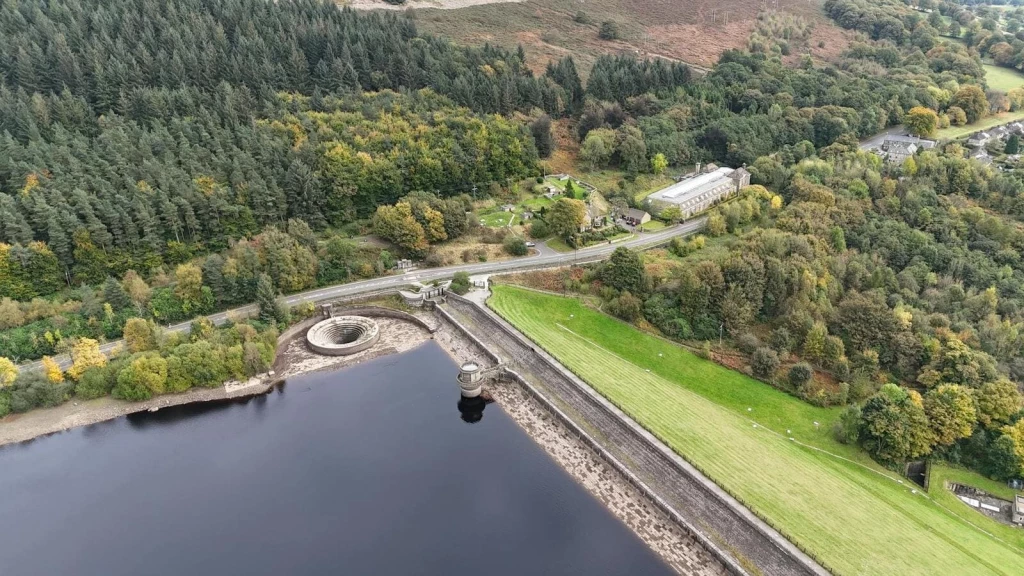 A scenic aerial view showcasing a reservoir surrounded by lush greenery and woodland, reflecting a Green Belt landscape offering opportunities for sustainable development under the new NPPF 2024. The image features a dam structure, an overflow spillway, and adjacent open fields, highlighting the potential for carefully planned growth while maintaining the area's natural character.