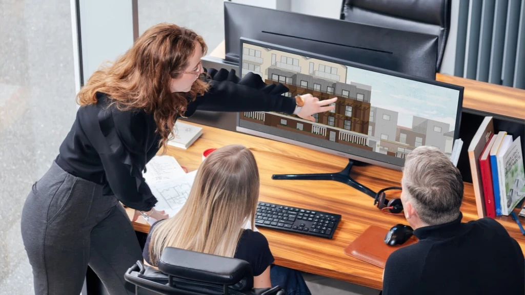 Architect presenting a detailed computer rendering of a proposed multi-storey residential development to colleagues, pointing out façade, window and upper-floor design features while architectural plans are spread across the desk.