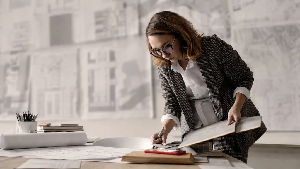 Female architect working at a studio desk, reviewing architectural drawings, floor plans and building material samples, with large technical plans and sketches displayed on the wall behind her.
