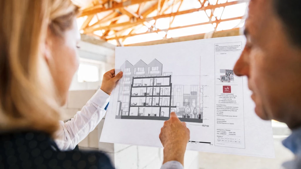 Architect and client reviewing a detailed architectural section drawing for a multi-storey residential development, pointing to proposed internal layouts and building levels while standing beneath exposed timber roof trusses on site.
