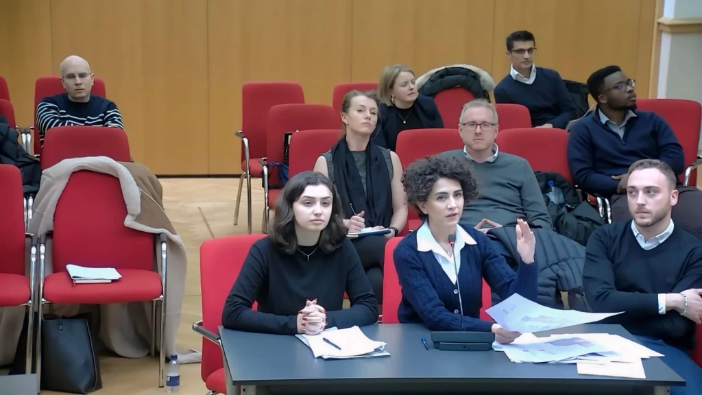 Planning consultant speaking into a microphone and presenting documents during a planning committee meeting, with architects, planning officers and other attendees seated in a council chamber listening to the discussion.