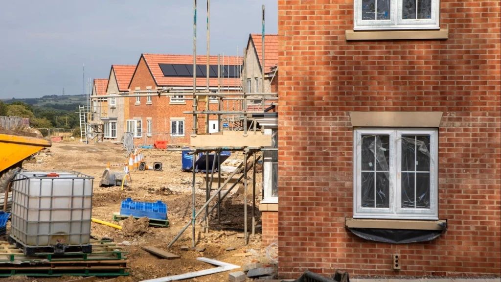 Residential housing development under construction, showing newly built red-brick homes, scaffolding, exposed groundworks and construction materials, with pitched tiled roofs and solar panels visible on completed sections.