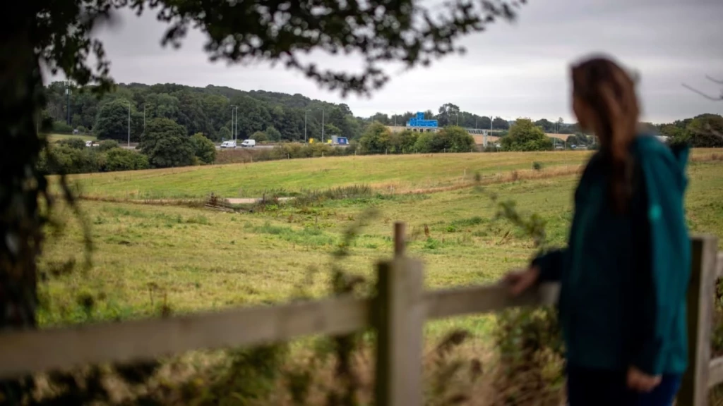 Open field beside a main road in the UK, viewed from behind a wooden fence with a person in the foreground.