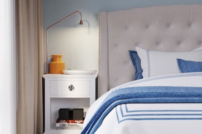 Stylish modern hotel bedroom detail featuring a tufted headboard, white nightstand with books, a trendy copper lamp, and bed linens with blue accents in a cosy room setting.