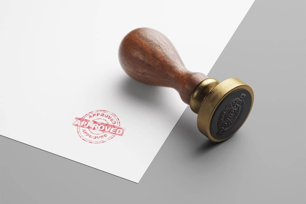 Close-up of a wooden stamp with a metal rim on a gray surface, leaving an 'Approved' imprint on a white document, representing the approval of planning permission for a development project.