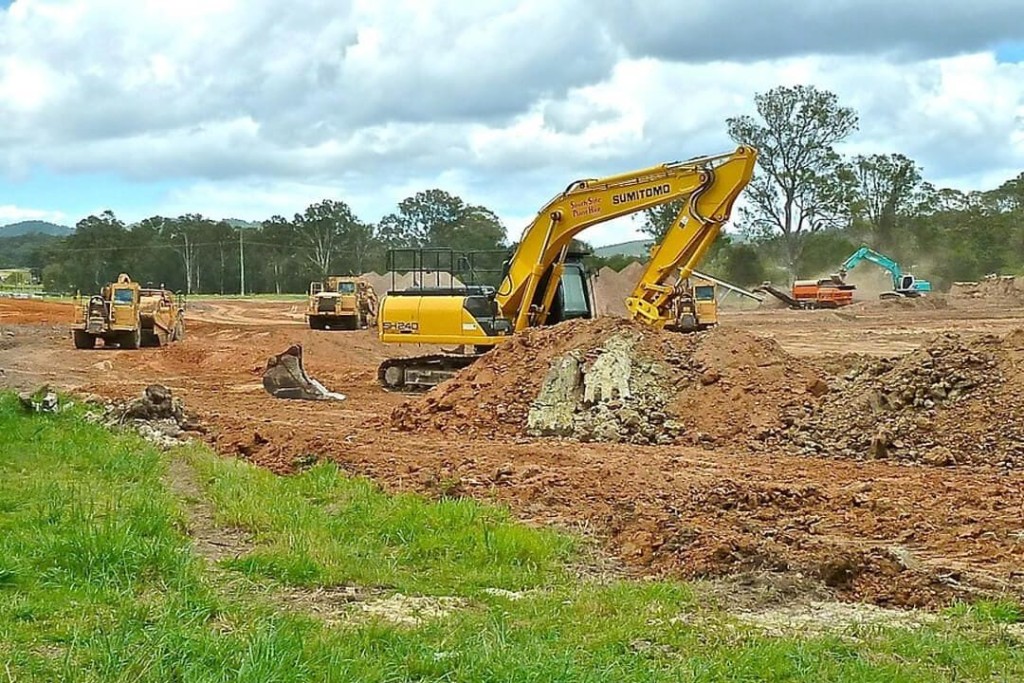 Construction machinery actively working on a development site with excavators and bulldozers preparing the land, surrounded by greenery and trees under a partly cloudy sky. 