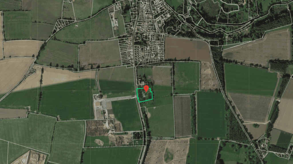 Overhead satellite view of a rural landscape with agricultural fields, featuring a red pinpoint marker on a highlighted green area, adjacent to a small development, illustrating land surveying or potential development planning.