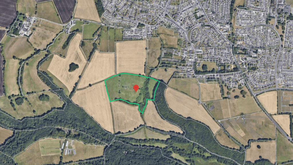 Satellite image showing a mix of urban and rural landscapes with a red location marker on a green outlined field, contrasting with surrounding developed areas, illustrating land use planning.