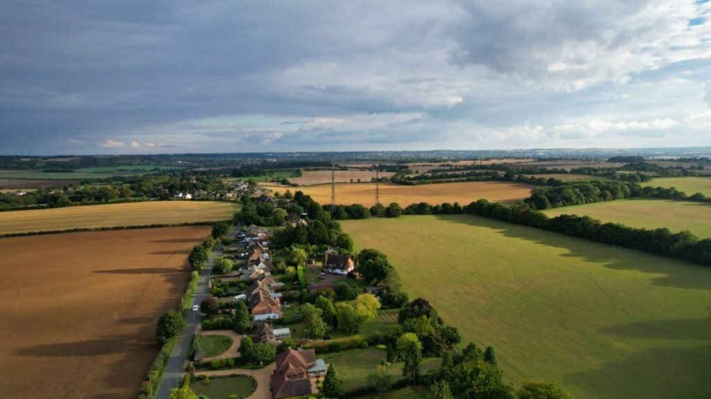Aerial view of a quaint English village nestled between lush green fields and golden agricultural land, with expansive views of the countryside under a dynamic sky, illustrating rural beauty in the England.