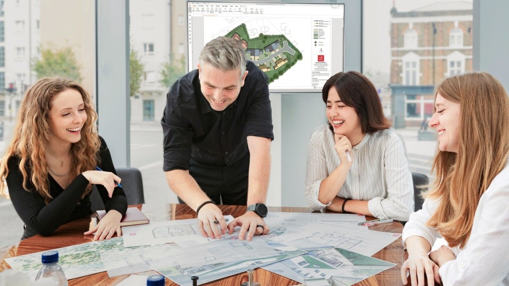 Enthusiastic team of urban planners engaged in collaborative discussion over architectural plans, with a computer screen displaying 3D housing project models in the background, in a bright office environment.