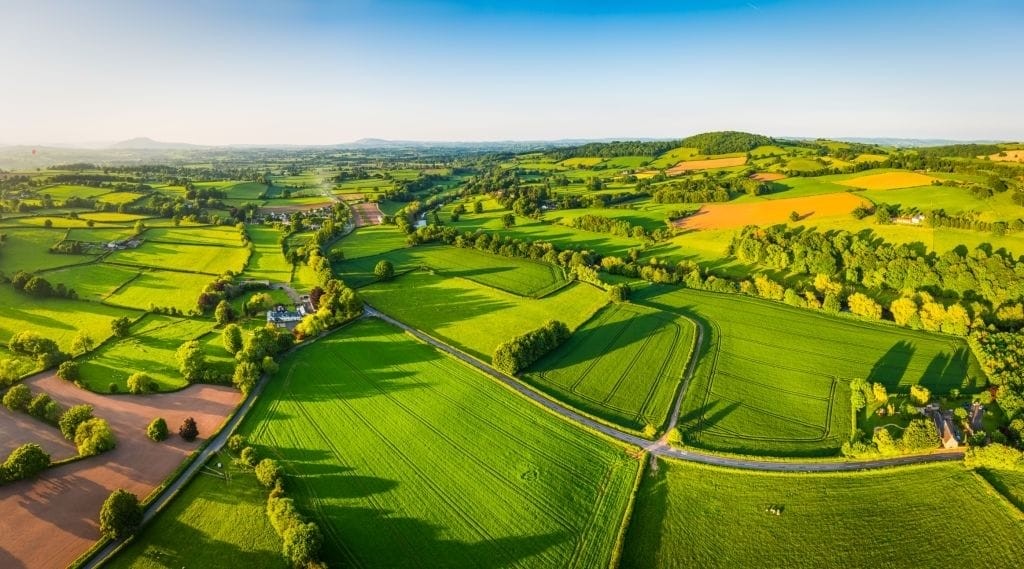 Aerial view of lush green fields and patchwork farmland in the UK countryside, with roads intersecting under a clear blue sky on a sunny day, highlighting the rural landscape's natural beauty and tranquility.
