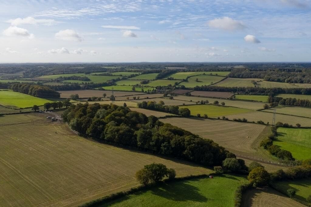 Aerial view of the lush English countryside, showcasing a patchwork of various agricultural fields, hedgerows, and clusters of trees under a partly cloudy sky, conveying a serene rural landscape.
