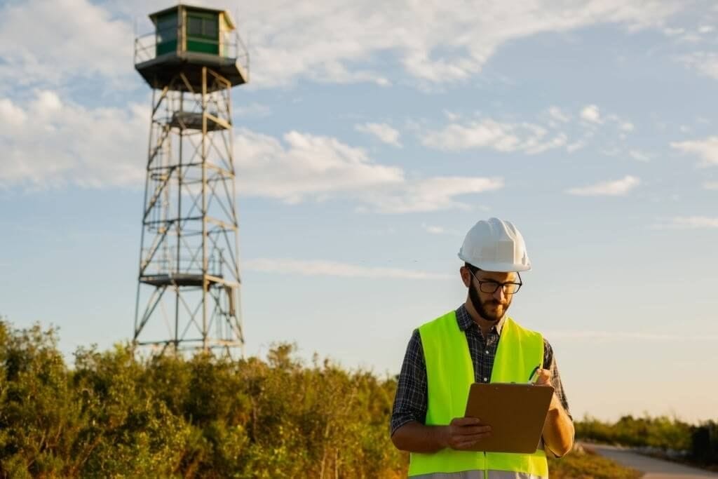 A focused engineer in a white hard hat and reflective vest taking notes on a clipboard in front of a tall observation tower with a green lookout post, set against a backdrop of a clear blue sky.