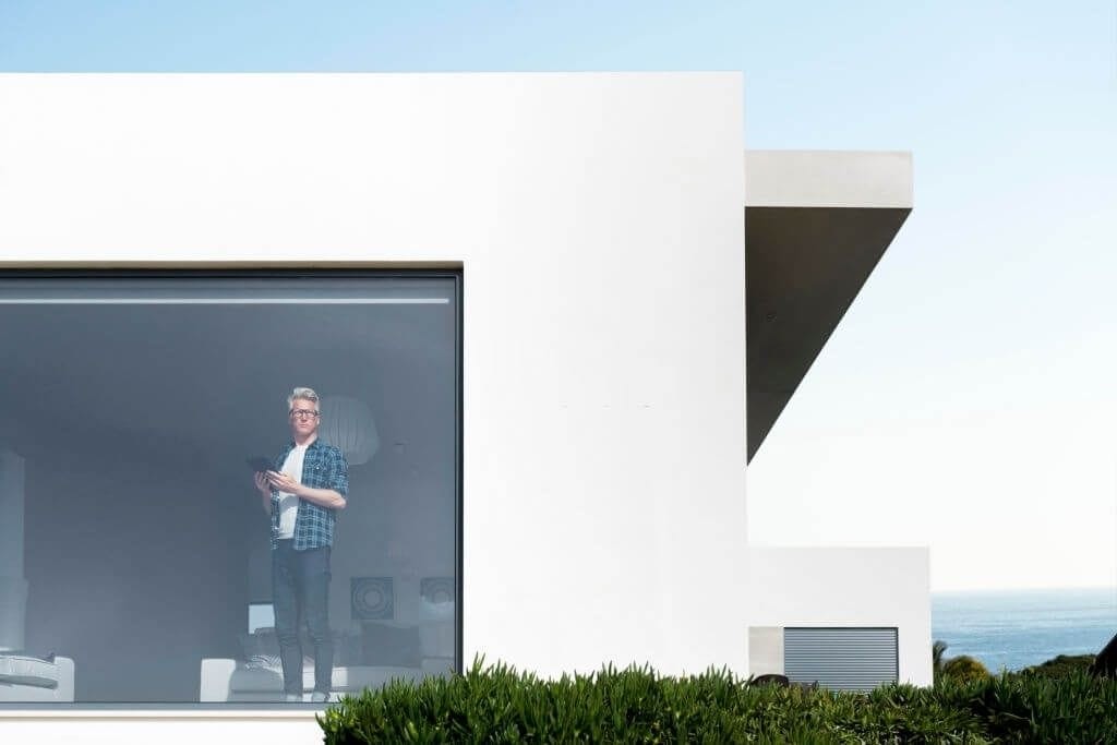 Man standing in the doorway of a modern minimalist white villa with large glass windows overlooking the ocean, under a clear blue sky, exemplifying contemporary architecture and luxury coastal living.