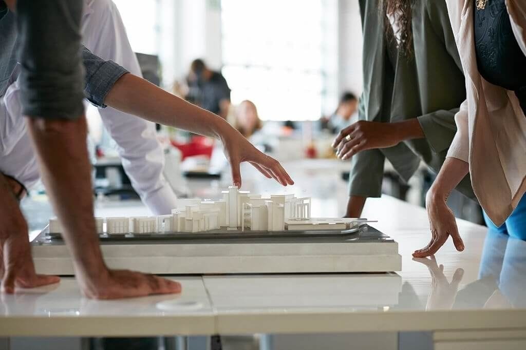 Close-up of professionals' hands gesturing over an architectural model of a building complex on a table, indicating discussion and planning in a brightly lit office environment.