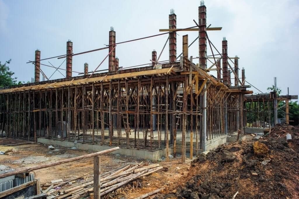 Early construction phase of a building with wooden scaffolding and concrete pillars on a construction site, showing the foundational framework and the beginning of a new structure under a cloudy sky.