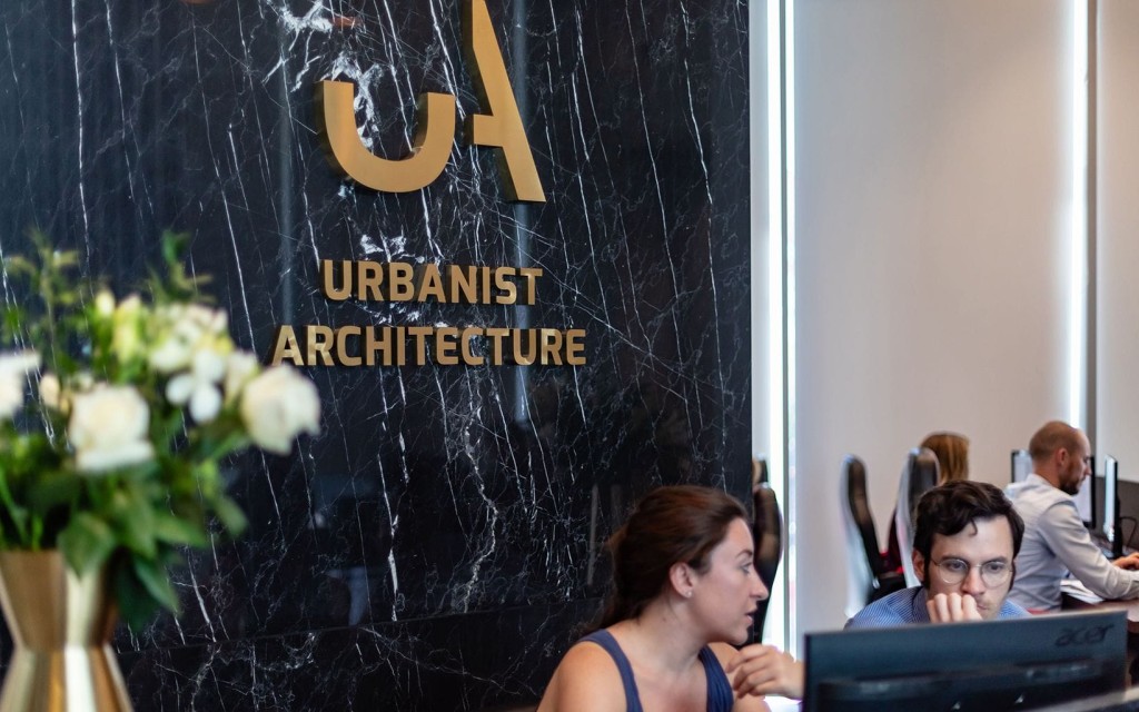 Interior of an architectural firm, 'Urbanist Architecture', with the company's gold logo on a black marble wall; professionals working diligently on computers in the background, depicting a busy, creative office environment.