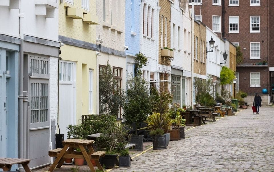 Quaint cobbled street lined with colorful mews houses, each adorned with potted plants and benches, in a serene urban residential area, portraying classic British charm and community-oriented living space.