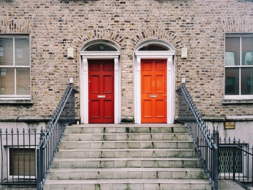 Symmetrical front view of a Georgian style brick townhouse with two vibrant doors, red and orange, each atop a set of stone stairs with wrought-iron railings, representing classic urban residential architecture in the UK.