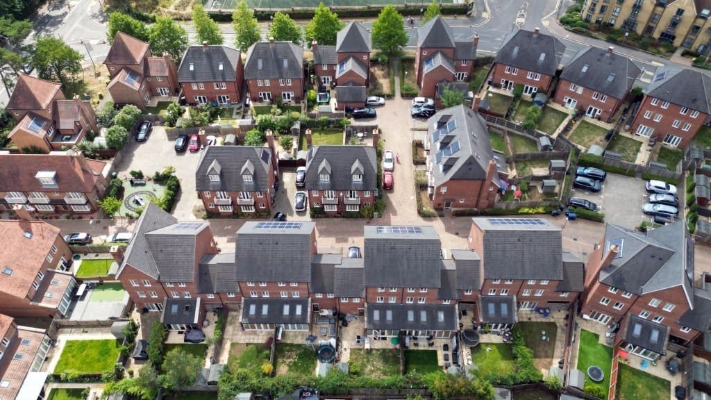 Aerial view of a suburban neighborhood in London showcasing a pattern of uniform red-brick houses with solar panels, well-kept backyards, and private driveways, illustrating modern residential planning.