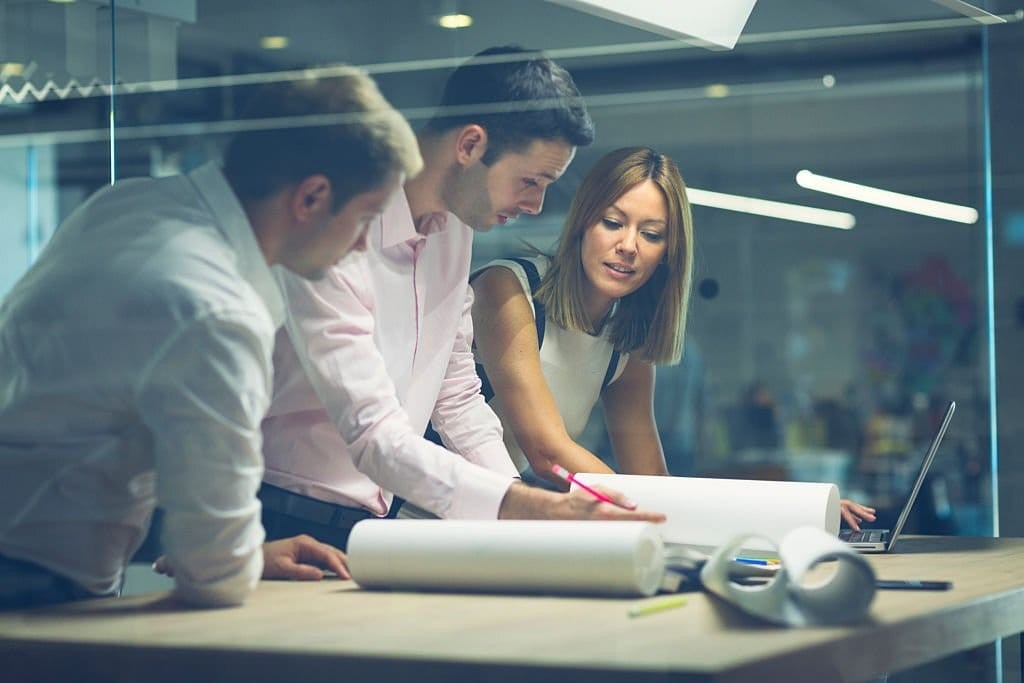 Three professional architects collaborating over a blueprint in a modern office environment, with focus on teamwork and innovative design planning.