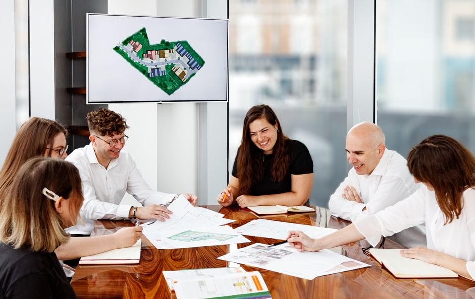 A diverse group of planning consultants and architects enthusiastically discussing project plans around a table, with site plans and a digital display showing a colourful aerial view of a development site in the background, in a modern office setting with large windows.