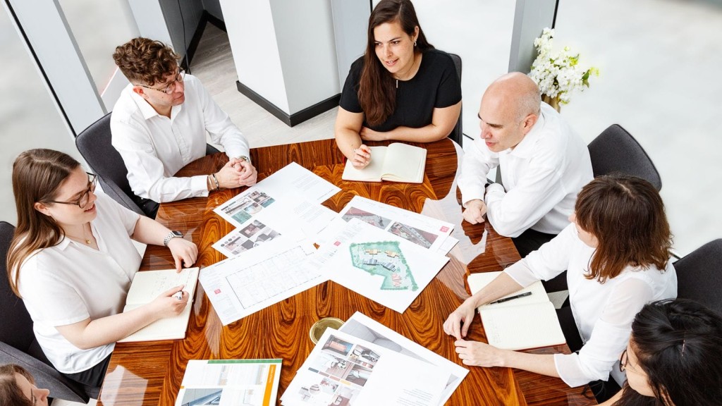 Overhead view of a professional meeting with a team of architects and planning consultants, engaged in discussion over a table scattered with various project documents, including maps, site plans, and architectural visuals, in a bright office environment.