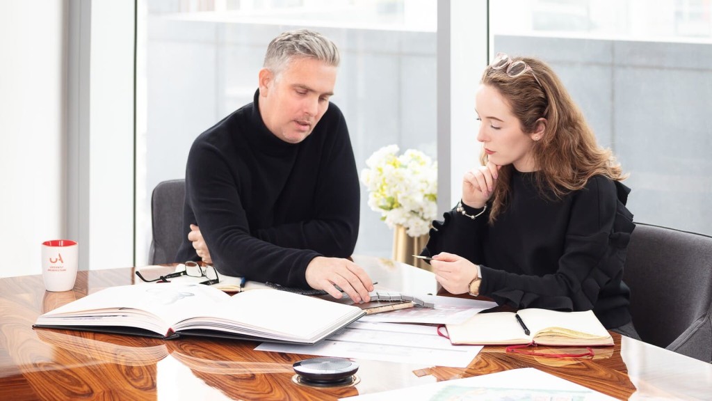 A professional meeting of two architects engaged in a discussion over a table scattered with various project documents, including maps, site plans, and architectural visuals, in a bright office environment.