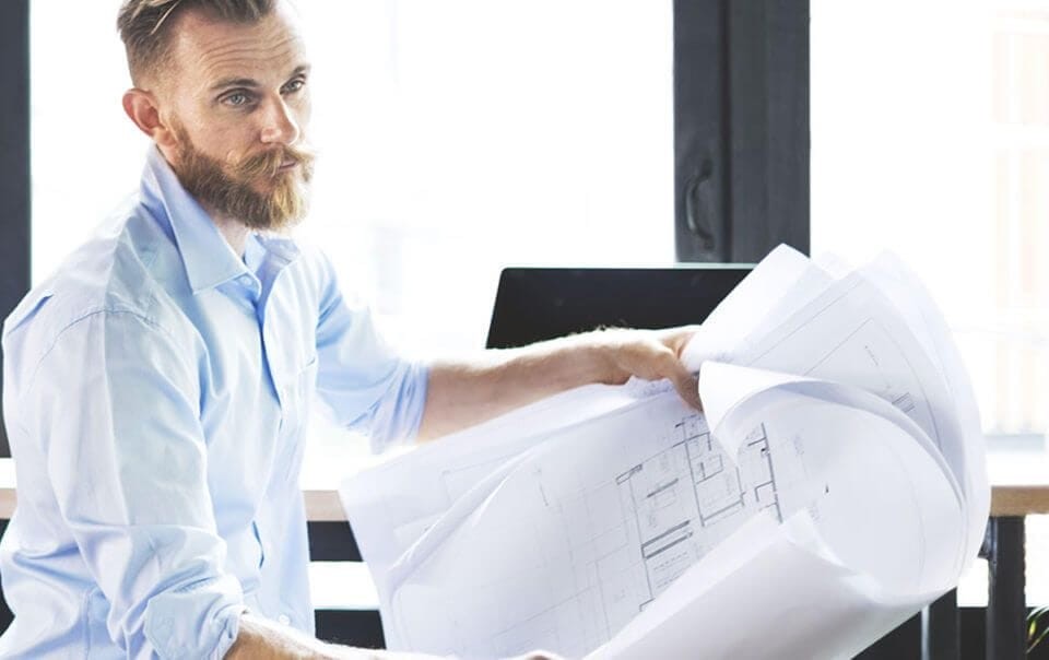 Focused male architect with a beard reviewing architectural blueprints in a well-lit office, symbolising detailed planning and design expertise in the architecture industry.