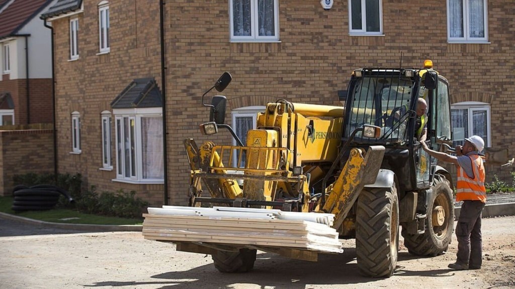 Construction workers in safety vests engaging in a discussion beside a yellow telescopic handler with building materials, in front of new brick houses on a residential street.