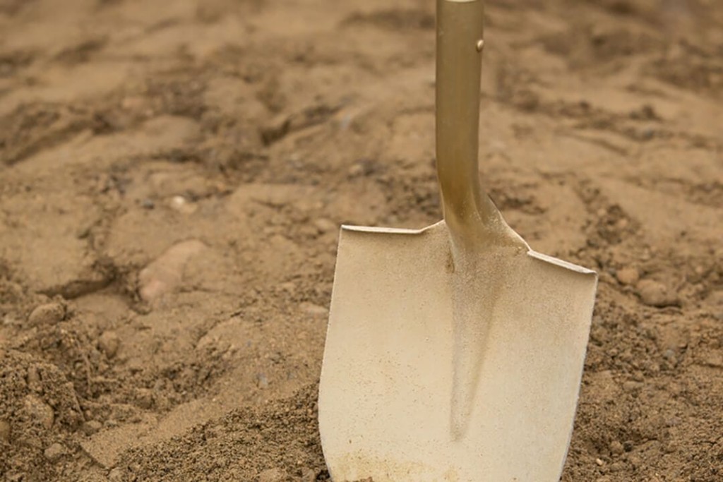Close-up of a shiny metal shovel embedded in sandy soil, indicating groundwork or the start of an excavation project.