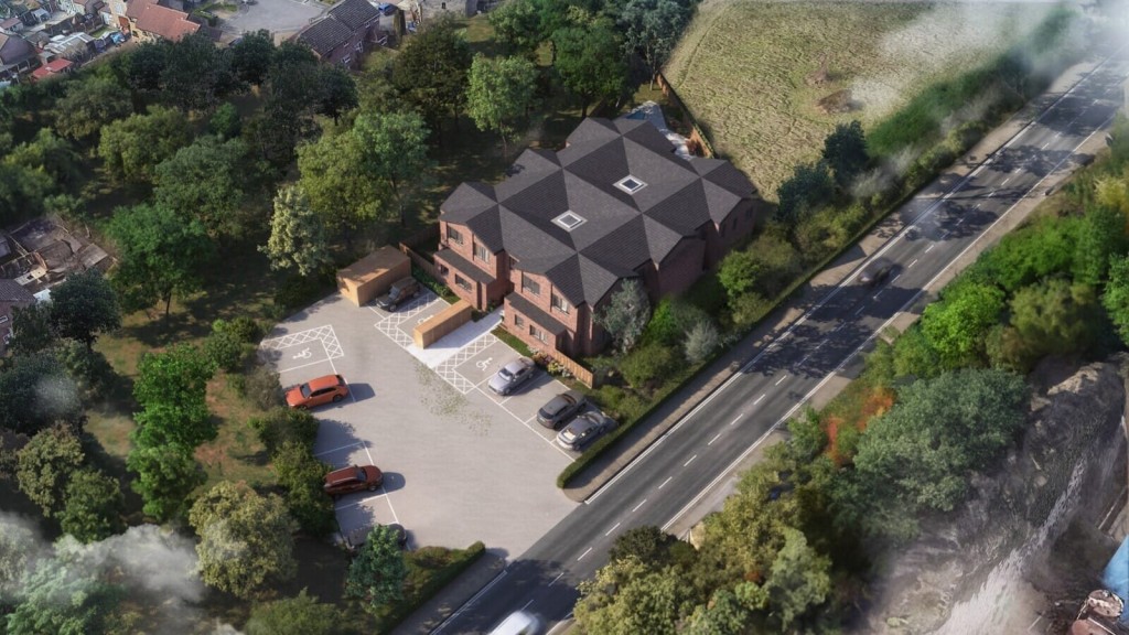 Aerial view of a new residential brick house with a dark shingle roof, surrounded by trees with a parking lot and adjacent road, showcasing suburban development.