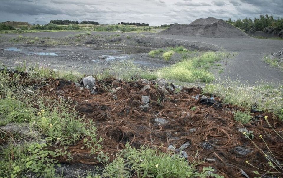 Discarded rusted metal wires strewn over brownfield land with overgrown vegetation and gravel piles under cloudy skies, depicting industrial neglect.