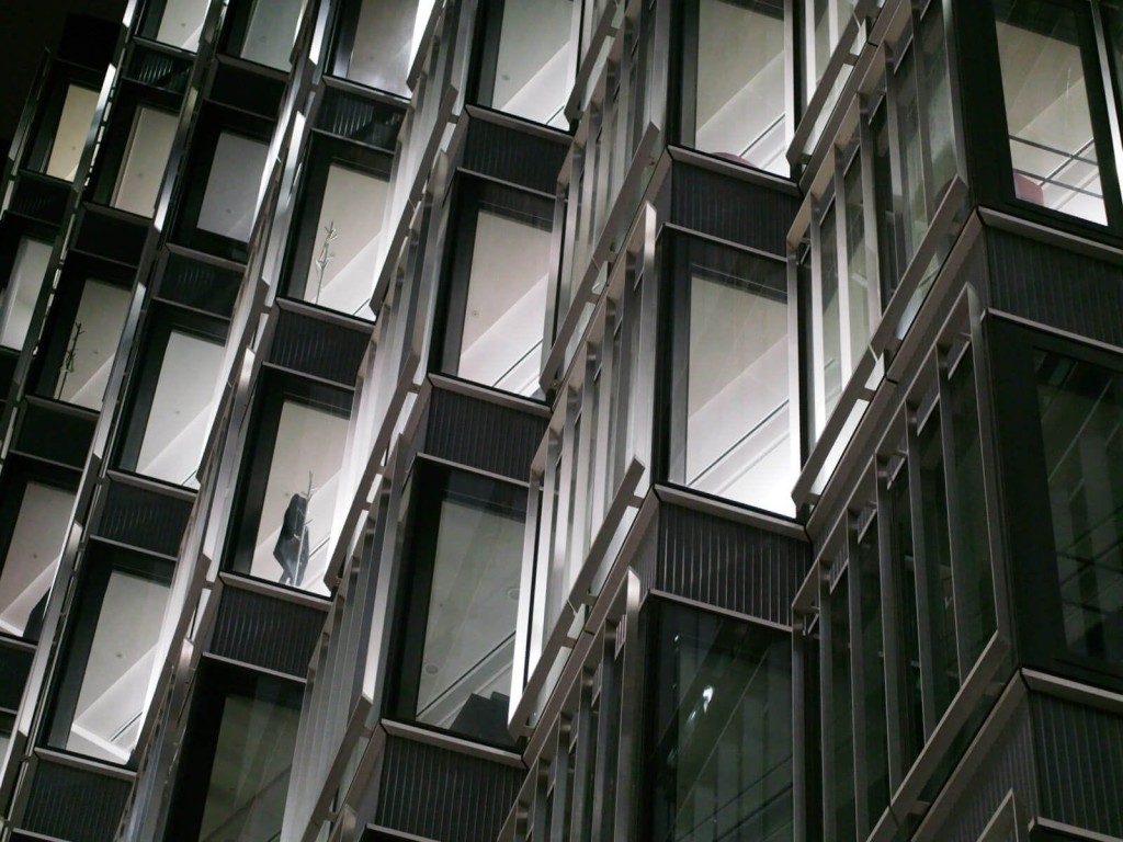 Close-up of a modern building facade at night, featuring geometric patterns of dark metal frames and large glass windows, reflecting the innovative design in contemporary architecture and urban development.